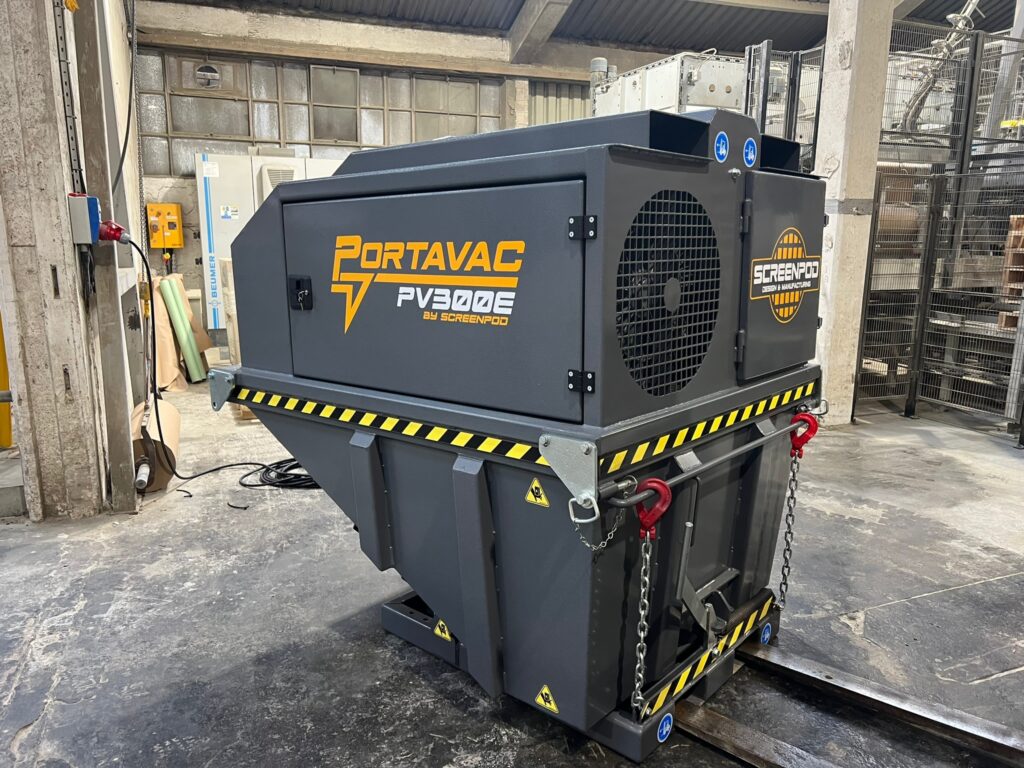 Ein dunkelgrauer Portavac PV300E von Screenpod steht in einer Werkhalle auf Schienen, mit gelb-schwarzen Warnstreifen, Ketten und roten Haken.