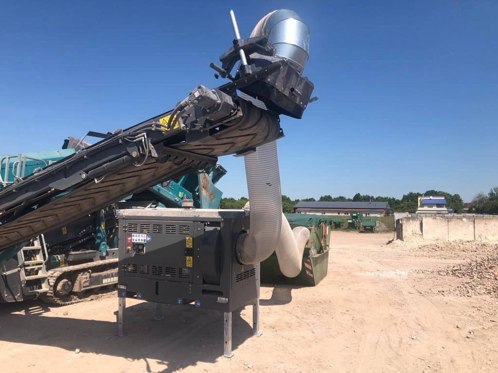 Unter wolkenlosem Himmel steht eine große Recyclingmaschine mit schwarzem Förderband, grauem Schlauch und silbernem Auswurf auf einem Schotterplatz.