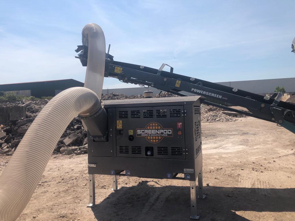 Ein metallischer Powerscreen-Förderer steht auf einem Schotterplatz neben einem Screenpod-Absaugmodul, darüber verlaufen zwei große flexible Schläuche.