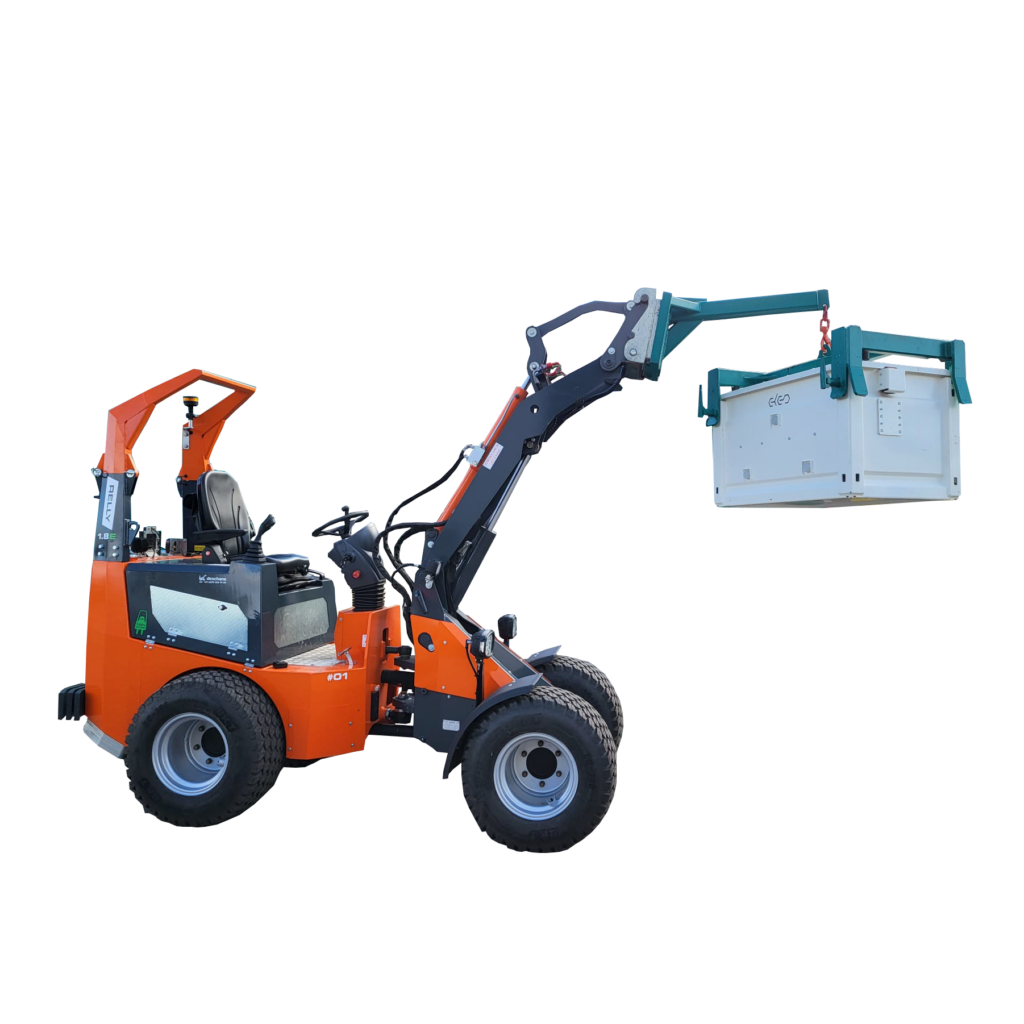 Der orange-schwarze Radlader RELLY 1.8E hebt in einem Hof einen weißen Container mit ausgefahrenem Hubarm neben einem grünen Anhänger und Gebäuden.