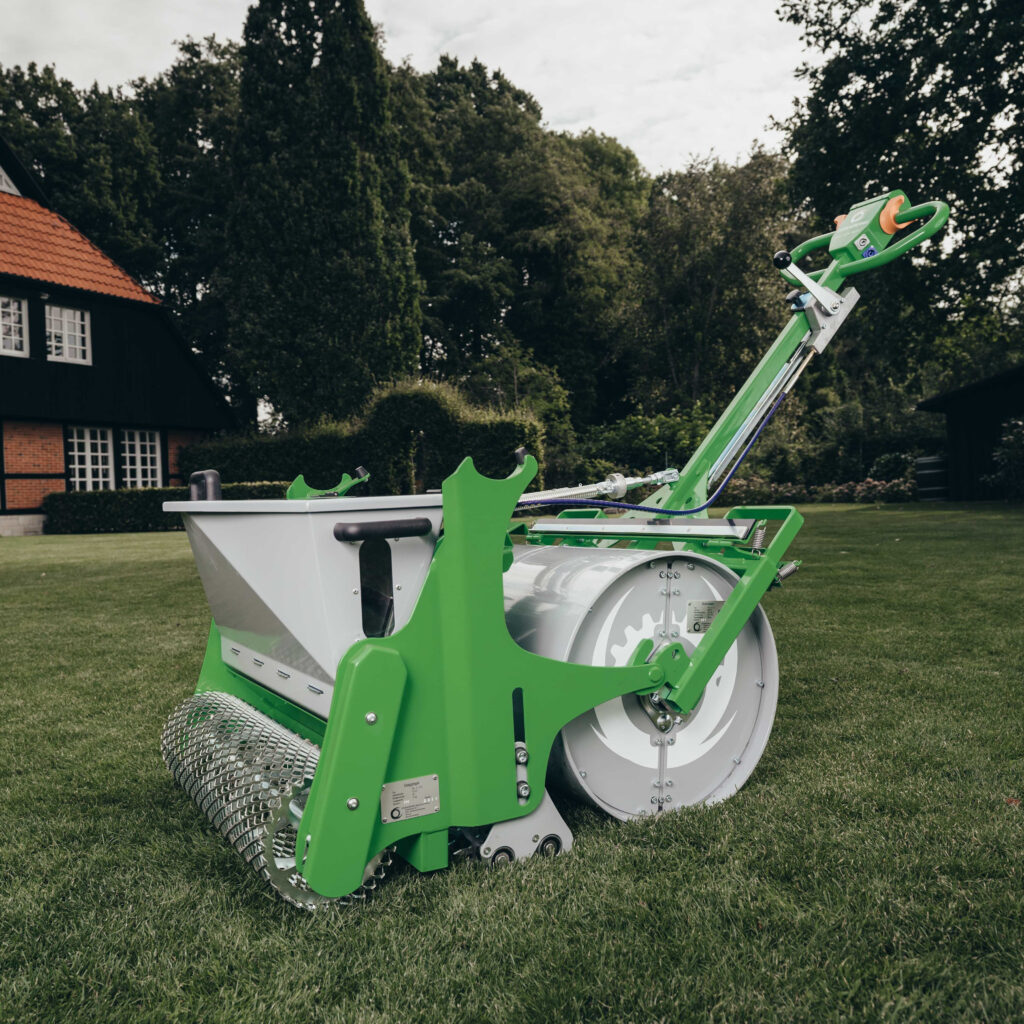 Ein grün-weißes Rasenbaumaschine-Modell steht auf einem gepflegten Rasen vor einem Haus mit rotem Dach und hohen Bäumen im Hintergrund.