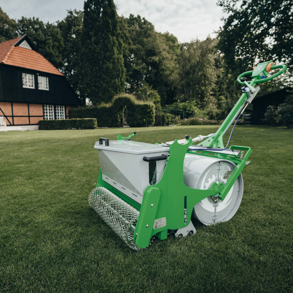 Eine grün-weiße Rasenbaumaschine steht auf einem gepflegten Rasen vor einem Fachwerkhaus, umgeben von hohen Bäumen im weitläufigen Garten.
