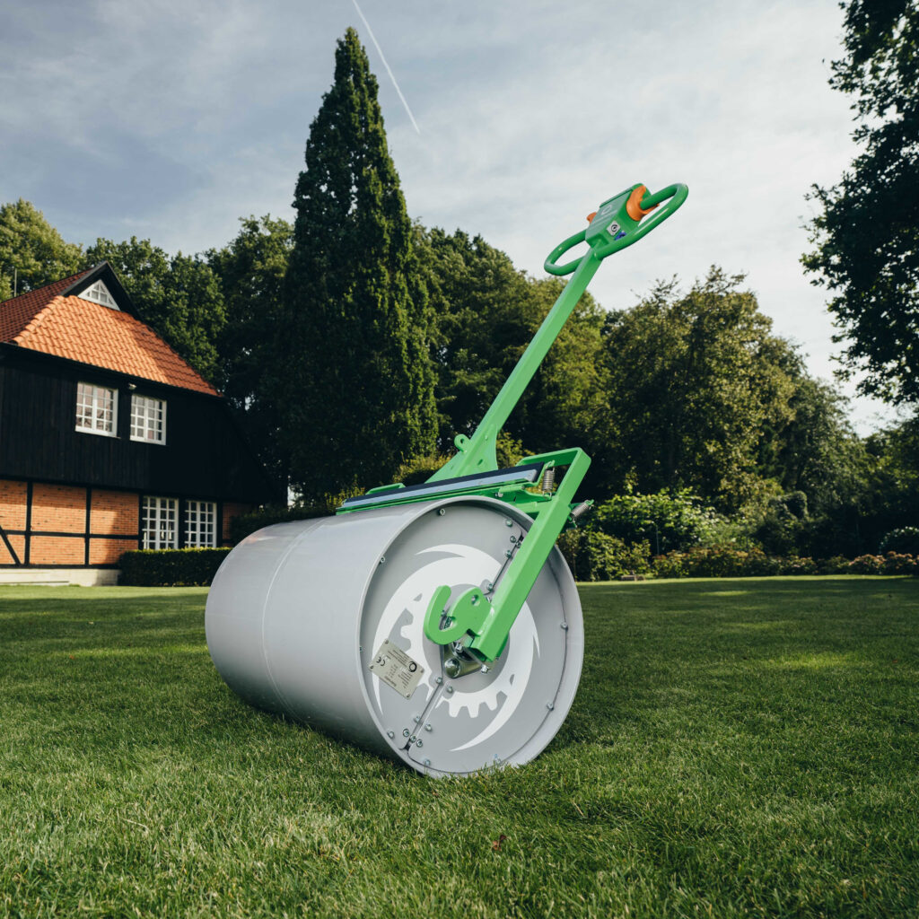 Ein grüner Rasenwalze mit langem Griff steht auf einem gepflegten Rasen vor einem Haus mit roten Ziegeldach und dichtem Baumbestand im Hintergrund.