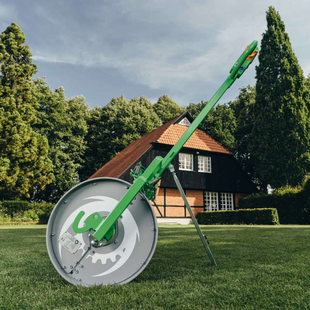 Eine grüne Rasenwalze mit langem Arm steht auf einem gepflegten Rasen vor einem Haus mit rotem Ziegeldach und hohen, dichten Bäumen im Garten.