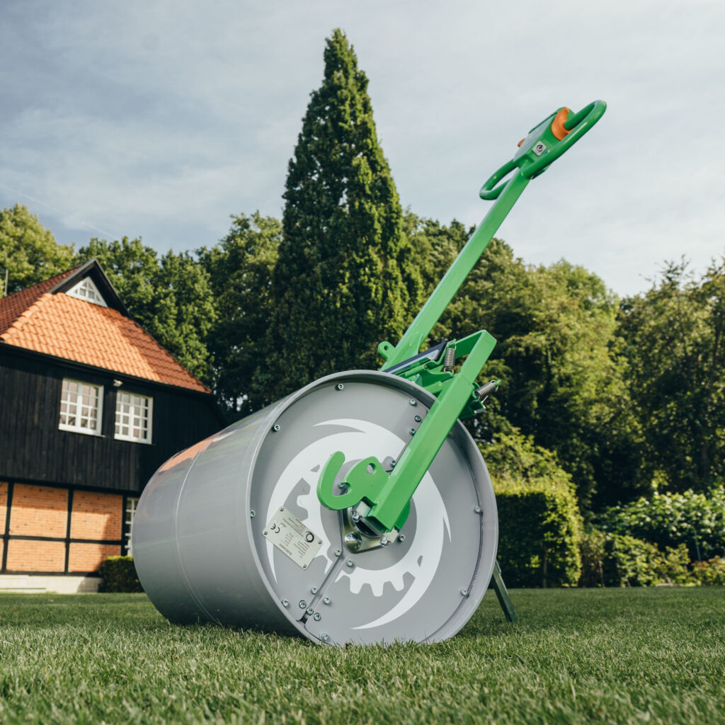 Eine grüne Rasenwalze mit silberner Trommel und langem Griff steht auf einer gepflegten Wiese vor einem Haus und hohen Bäumen im Garten.
