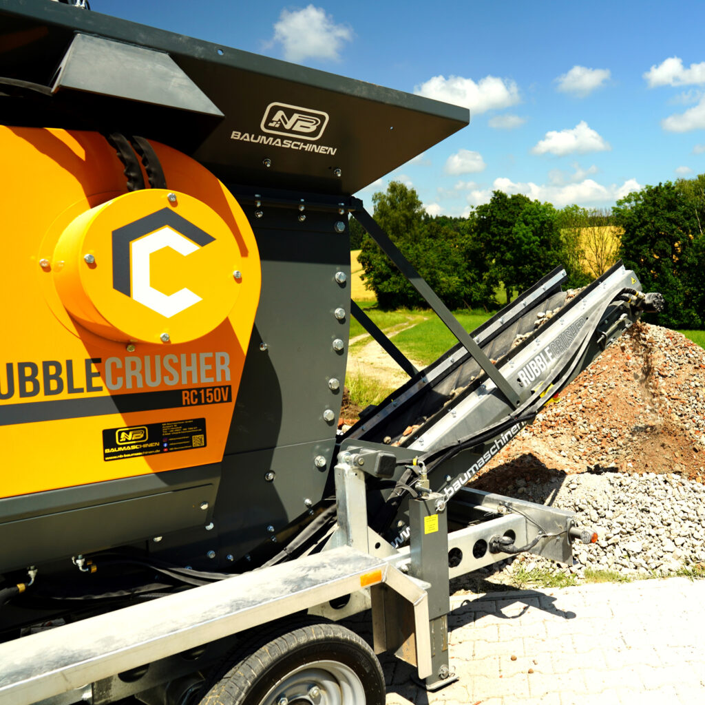 Der orange-schwarze Rubble Crusher RC150V fördert Schutt über ein Förderband, während im Hintergrund blauer Himmel und grüne Bäume zu sehen sind.