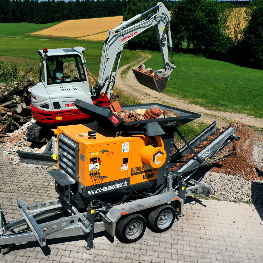 Auf einem Feldweg steht ein orange-schwarzer Rubble Crusher RC150 mit Kettenfahrwerk, während ein Bagger Schutt in den Trichter lädt.