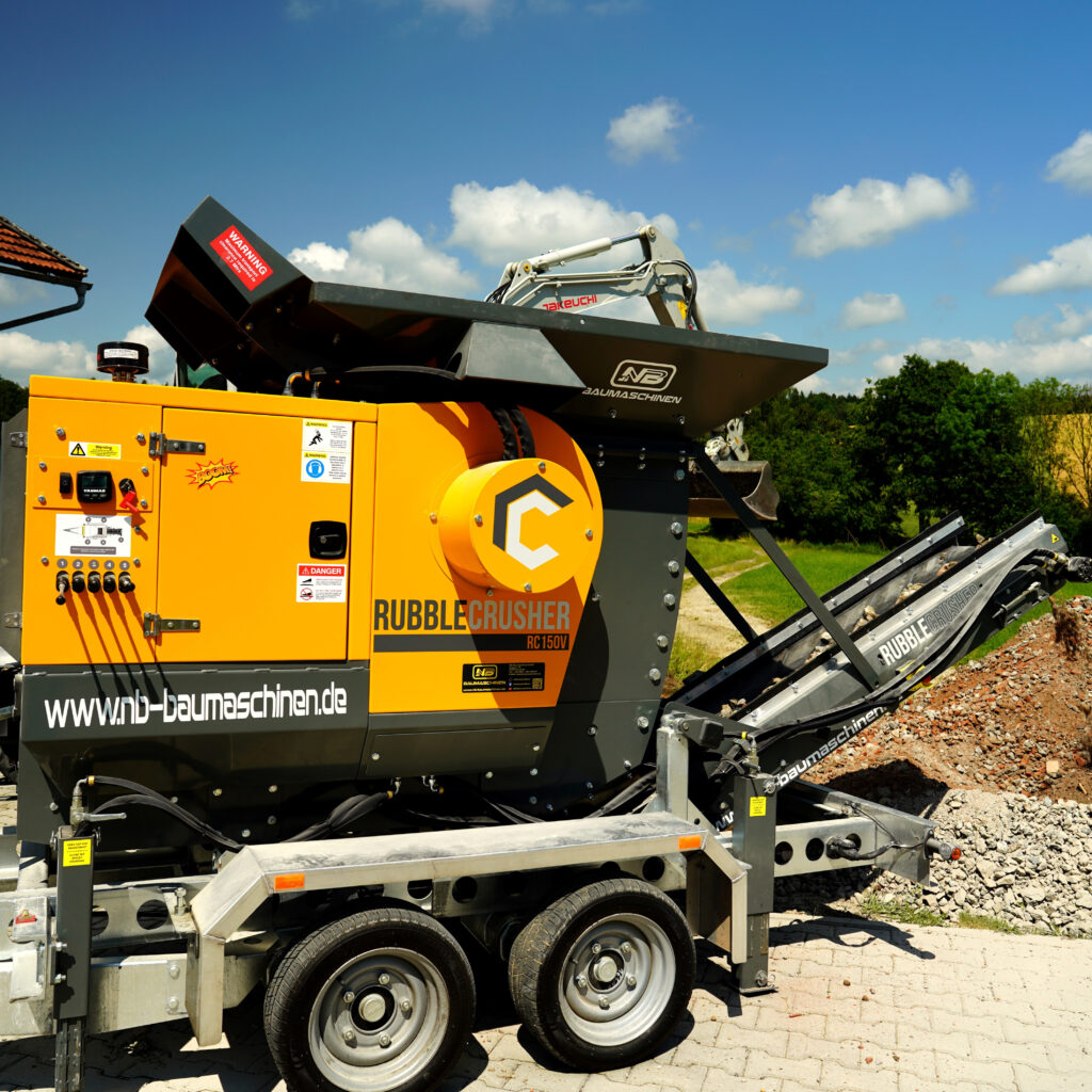Gelb-schwarzer Rubble Crusher RC150V auf Anhänger, daneben ein Takeuchi-Baggerarm und Förderband vor blauem Himmel mit weißen Wolken.