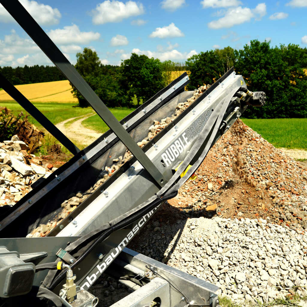 Ein schräg montierter Rubble Crusher RC150-4 fördert Bauschutt über ein Förderband in einen Steinhaufen vor grüner Landschaft und blauem Himmel.