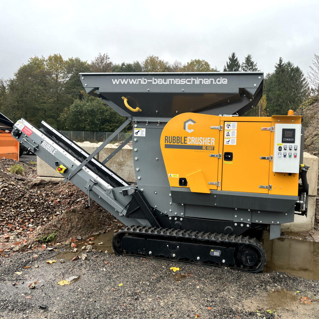 Ein orange-grauer RubbleCrusher RC150T mit Förderband und Steuerkasten steht auf einer nassen Baustellenfläche vor Bäumen.
