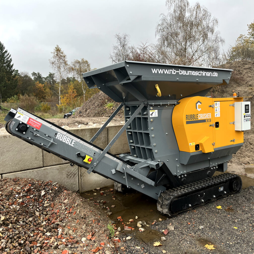 Große RubbleCrusher RC150T-2 Brechanlage in Grau und Gelb steht auf Kettenfahrwerk neben einem Schutt- und Erdwall, mit Förderband und Trichter.