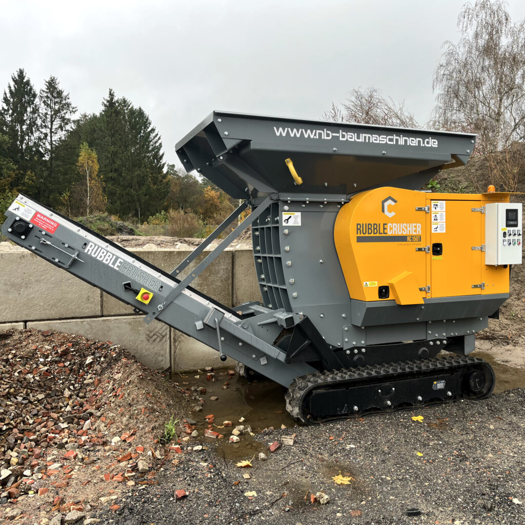 Der gelb-graue RubbleCrusher RC150T-3 steht auf Raupen neben Betonblöcken, mit großem Trichter, Förderband und Steuergehäuse vor bewaldetem Hang.
