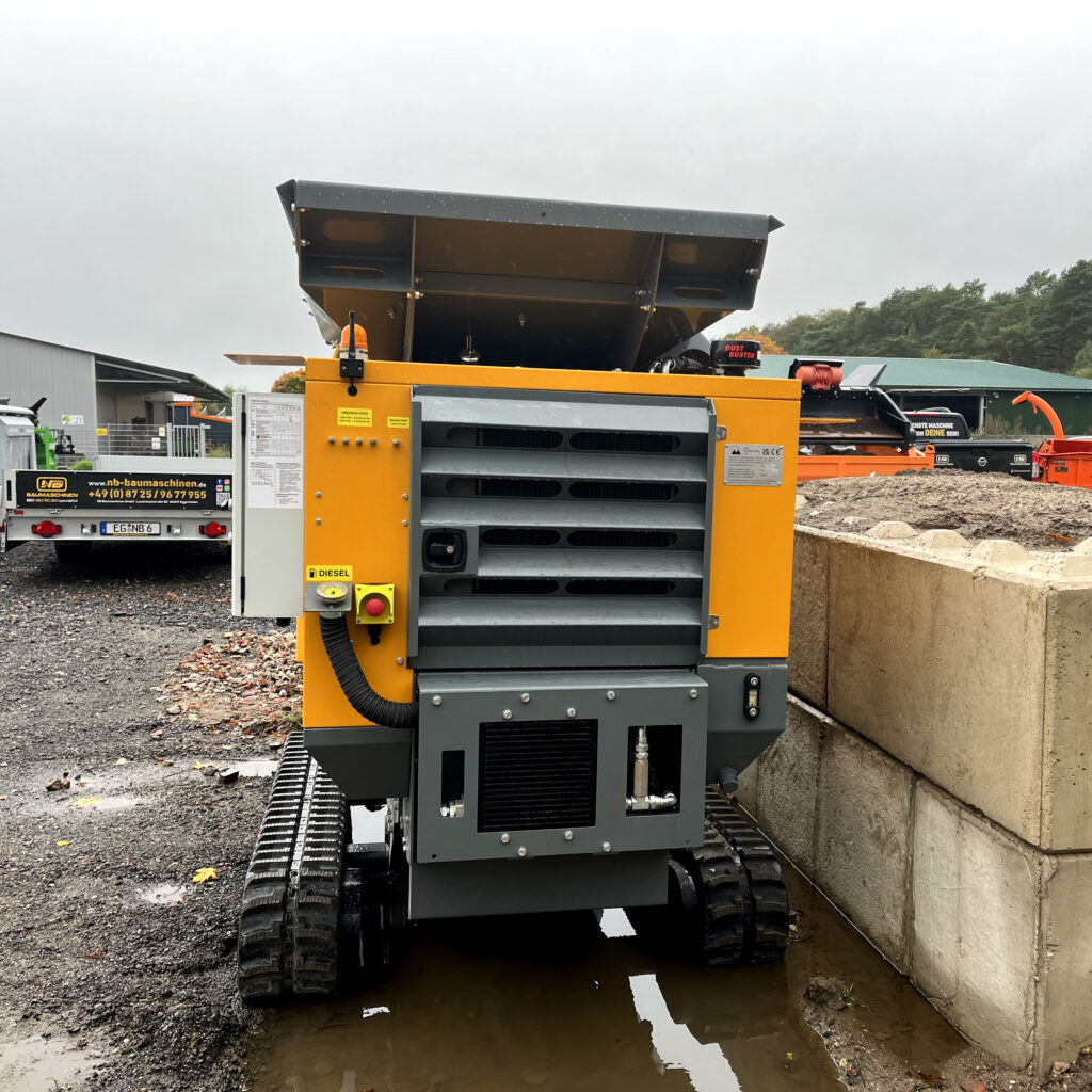 Der gelb-graue RubbleCrusher RC150T-5 steht auf Raupen neben einer Betonwand, mit großem Aufgabebunker, Dieselanschluss und Dust Buster.