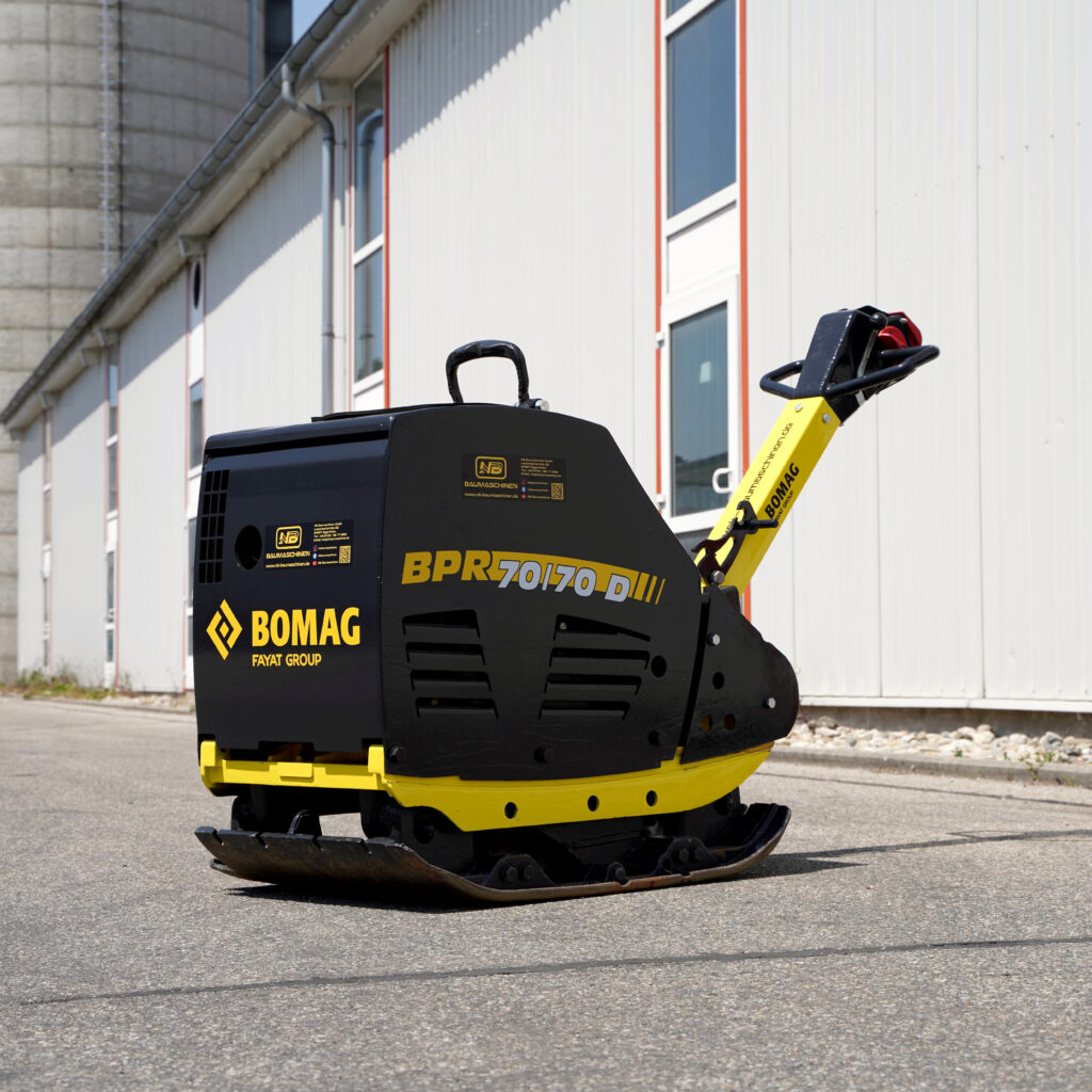 Schwarze und gelbe BOMAG Rüttelplatte BPR 70/70D mit Führungsbügel steht vor einer hellen Industriefassade mit schmalen Fenstern und Rohren.