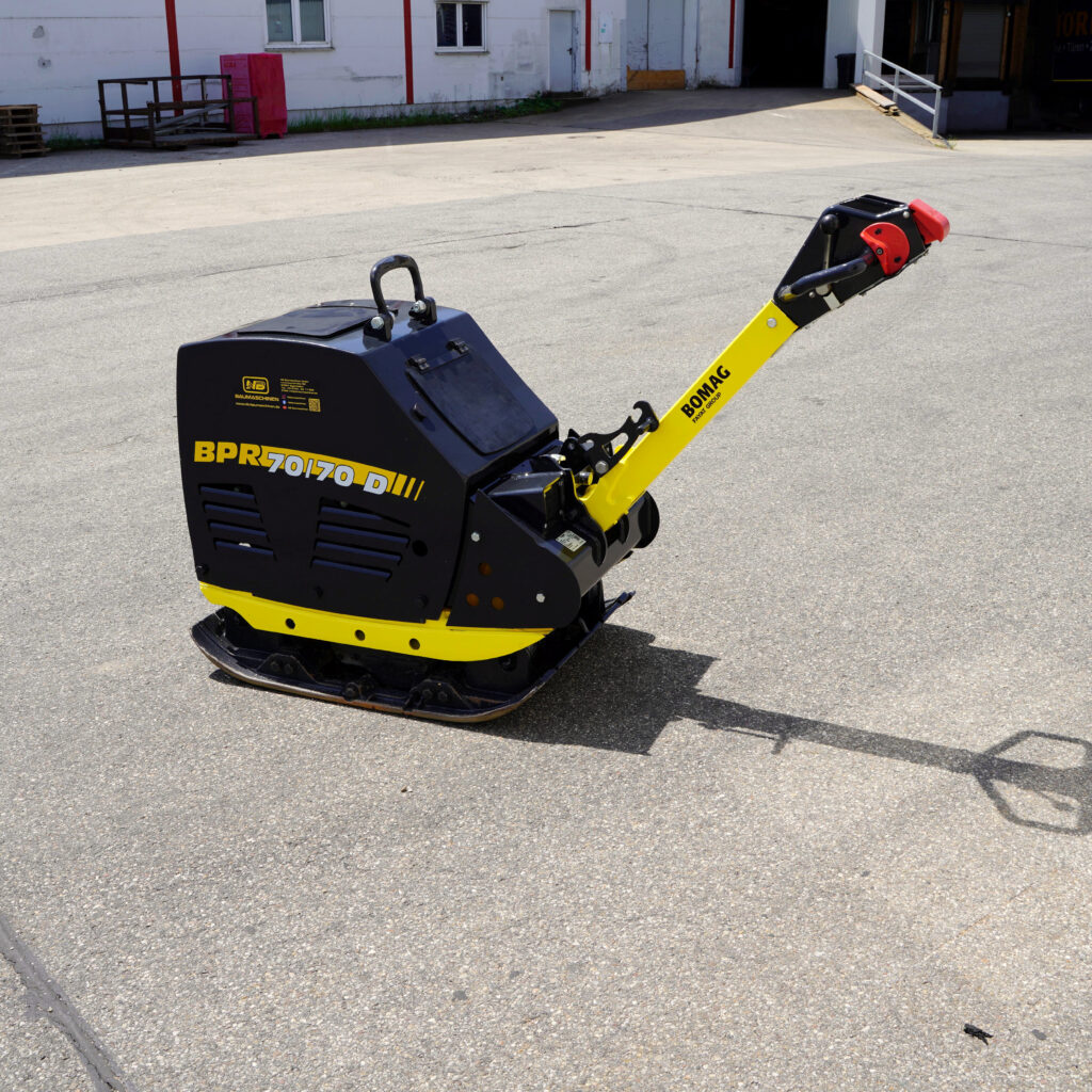Ein gelb-schwarzer BOMAG Rüttelplatte BPR 70/70 D steht auf Asphalt vor einem Industriegebäude, der lange Führungsgriff ragt nach rechts.