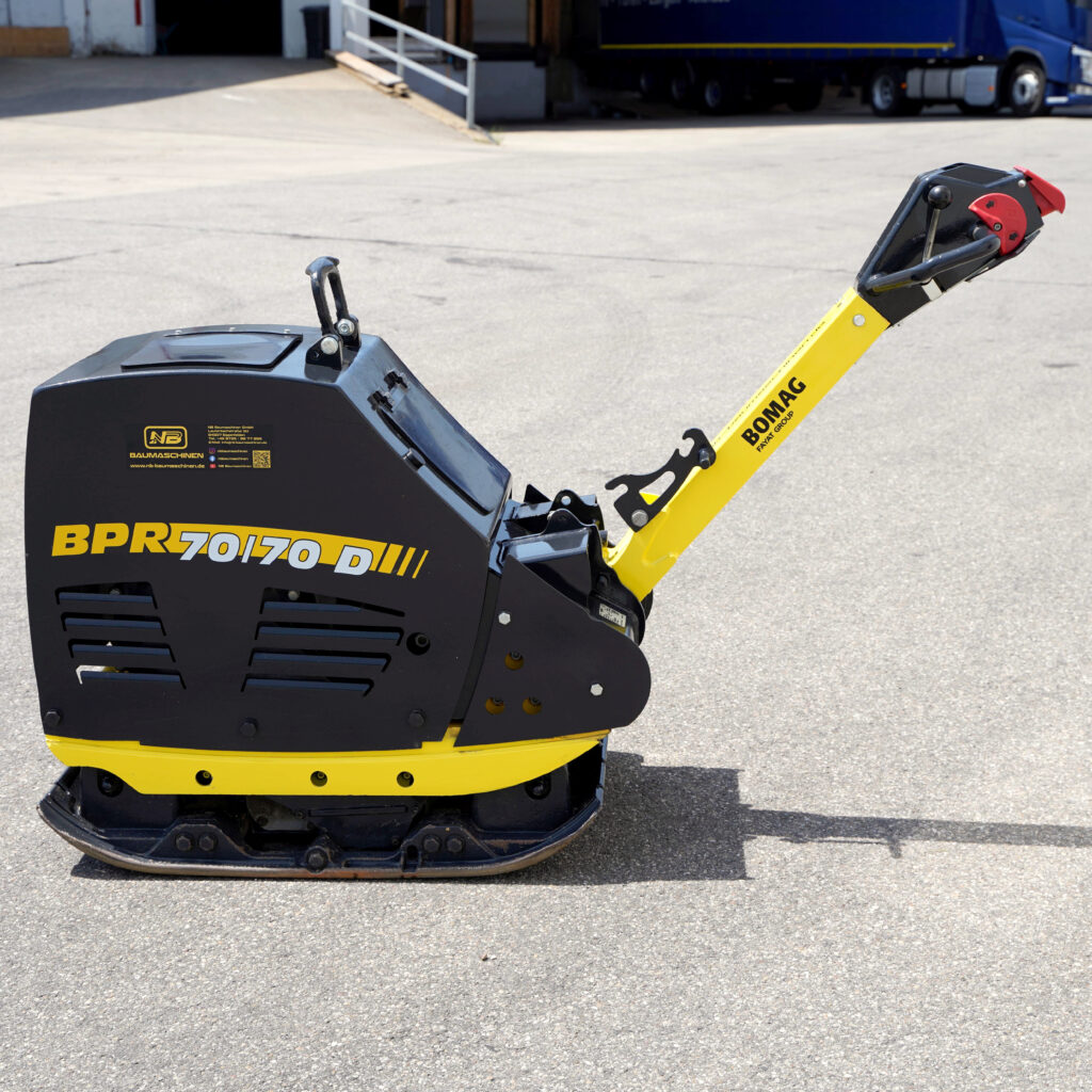 Schwarze und gelbe BOMAG Rüttelplatte BPR 70/70D-5 mit langem Führungsgriff steht auf Asphalt vor einem Industriegebäude im Tageslicht.