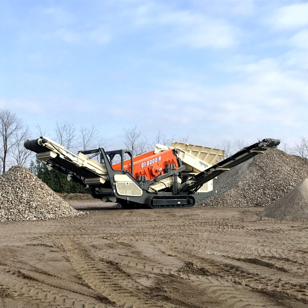 Eine orange-beige Siebanlage GI 5000 R steht zwischen großen Schotterhaufen auf einem staubigen Platz unter wolkigem Himmel mit ausgefahrenen Förderbändern.