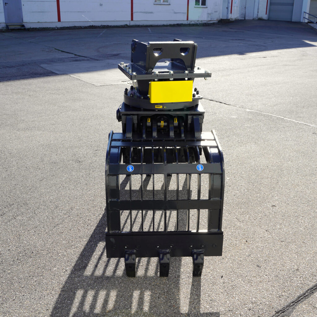 Ein schwarzer Sortiergreifer PG450-2 mit gelbem Gehäuse steht auf dem Asphalt vor einer Industriehalle im Sonnenlicht und zeigt seine stabilen Zinken.