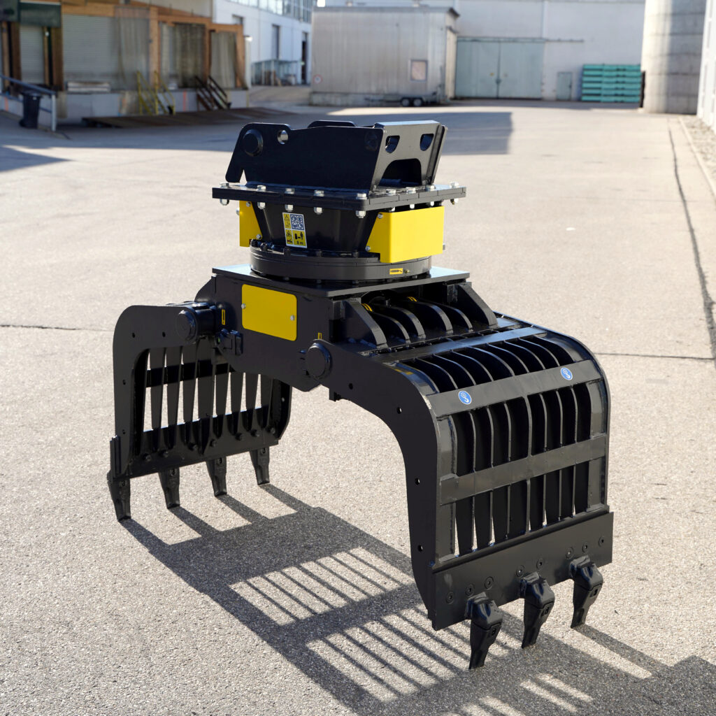 Ein schwarzer und gelber Sortiergreifer mit langen Zinken steht auf Asphalt vor Industriegebäuden im Sonnenlicht, mit klaren Schatten und breiter Aufnahme.