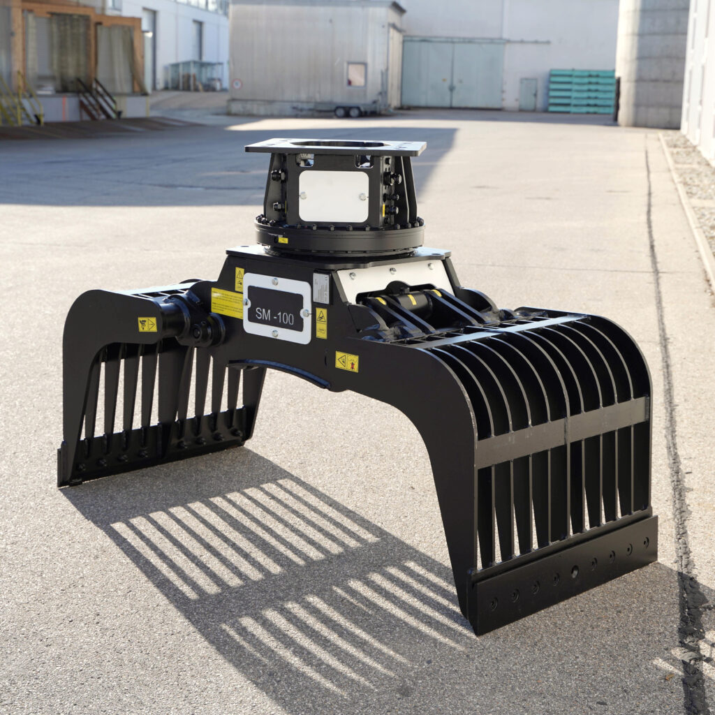 Ein schwarzer Sortiergreifer SM-100 mit breiten Greifzinken steht auf einem Asphaltplatz vor Industriegebäuden im hellen Sonnenlicht.
