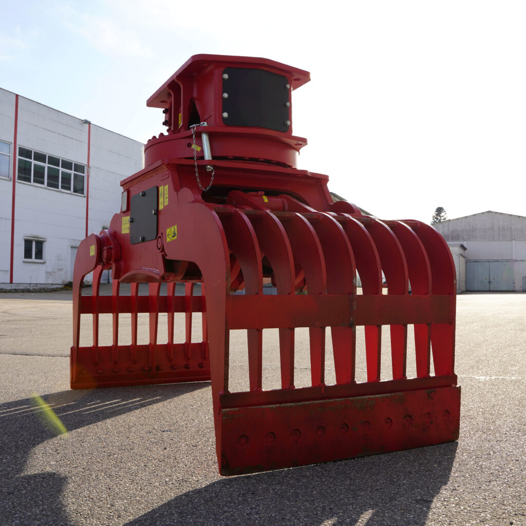 Massiver roter Sortiergreifer SM-120-1 mit breiten Zinken und schwarzer Oberseite steht auf einem Industriegelände vor hellen Hallen und Himmel.