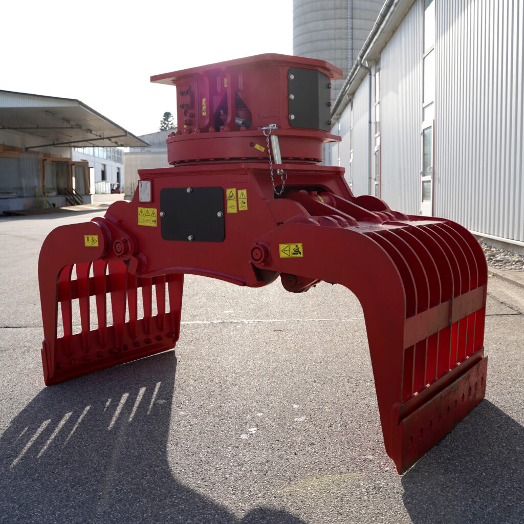 Ein großer roter Sortiergreifer mit breiten Greifarmen steht auf einem Hof vor Industriebauten und zeigt gelbe Warnhinweise sowie schwarze Wartungsklappen.