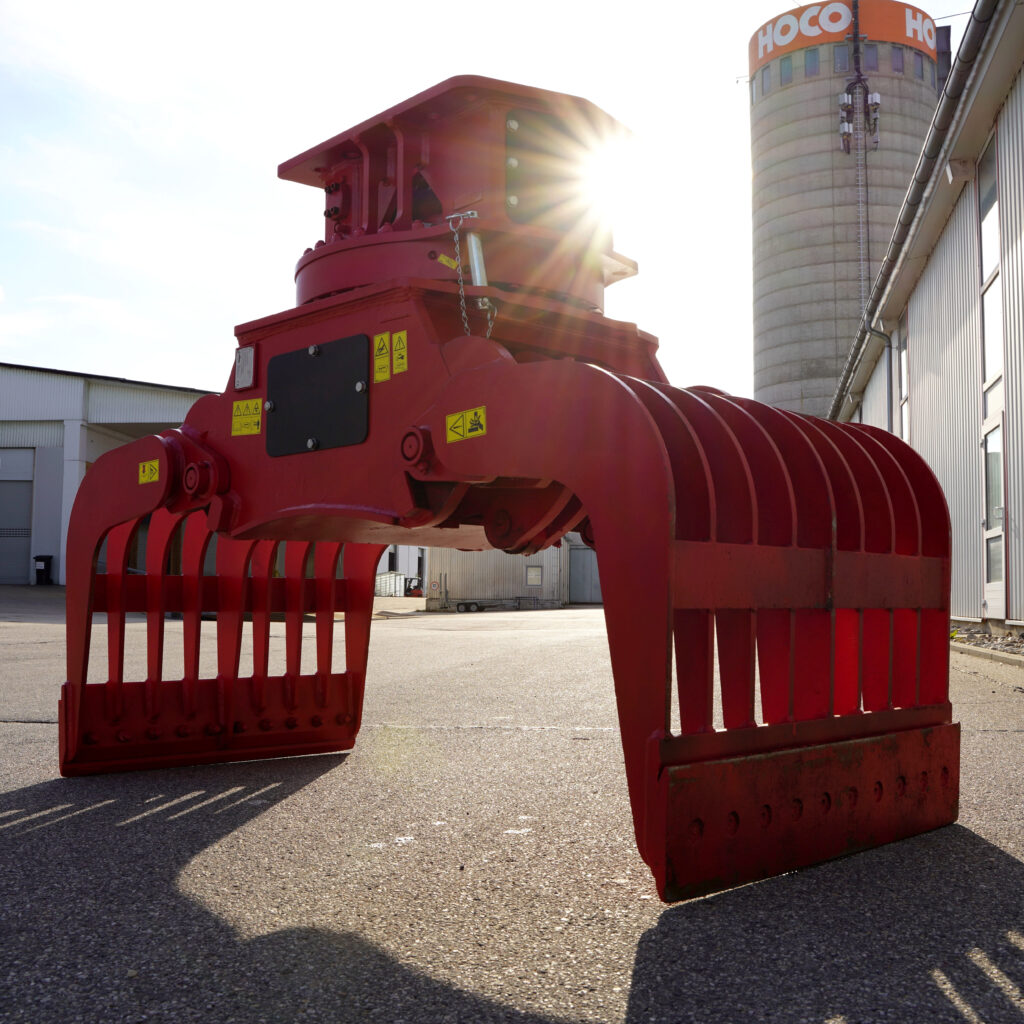 Ein roter Sortiergreifer mit breiten Zinken steht auf dem Hof vor Industriegebäuden, während Sonnenlicht durch die Konstruktion fällt.