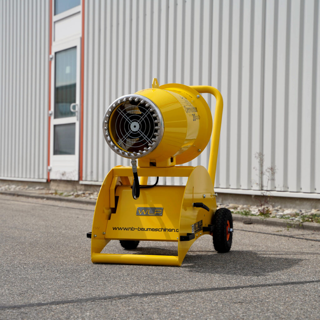 Eine gelbe Demolitor 20 Staubbindeanlage steht vor einer hellen Industriehalle, mit großem Ventilator, Griff und schwarzen Rädern.