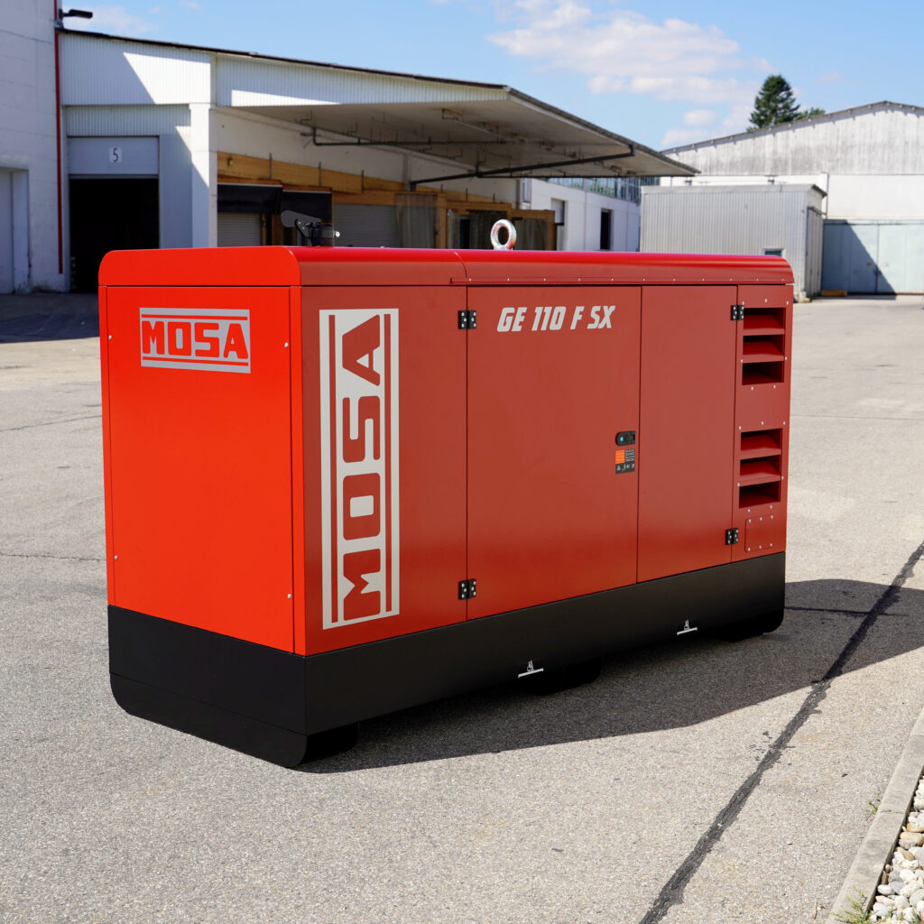 Roter Mosa GE 110 F SX Stromerzeuger mit schwarzem Sockel steht vor Industriehallen auf Asphalt, mit gut sichtbaren Türen, Lüftungsschlitzen und Hebeöse.