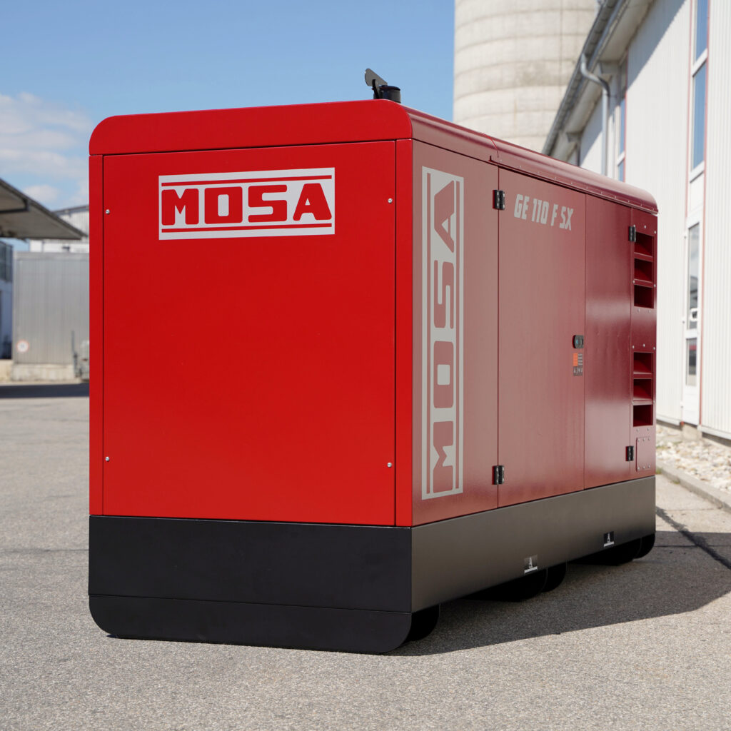 Ein roter, geschlossener MOSA GE 110 FSX Stromerzeuger steht vor einer Industriehalle, mit schwarzem Sockel und seitlichen Serviceklappen.
