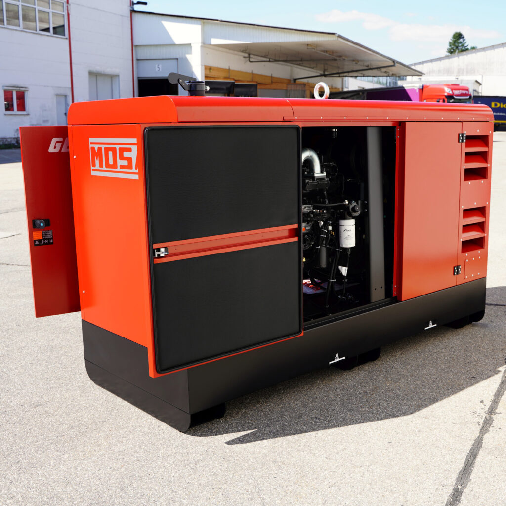 Ein roter, geschlossener Stromerzeuger mit schwarzer Seitenklappe steht vor einer Industriehalle; im Inneren sind Motorbauteile sichtbar.