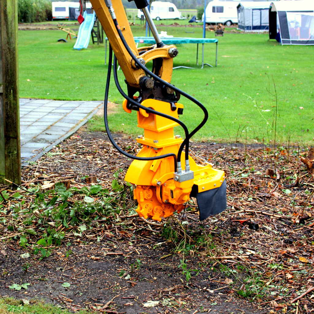 Ein gelber Stubbenfräse-Aufsatz am Bagger arbeitet an einem Baumstumpf auf einer feuchten Rasenfläche mit Holzschnitzeln und Garten im Hintergrund.