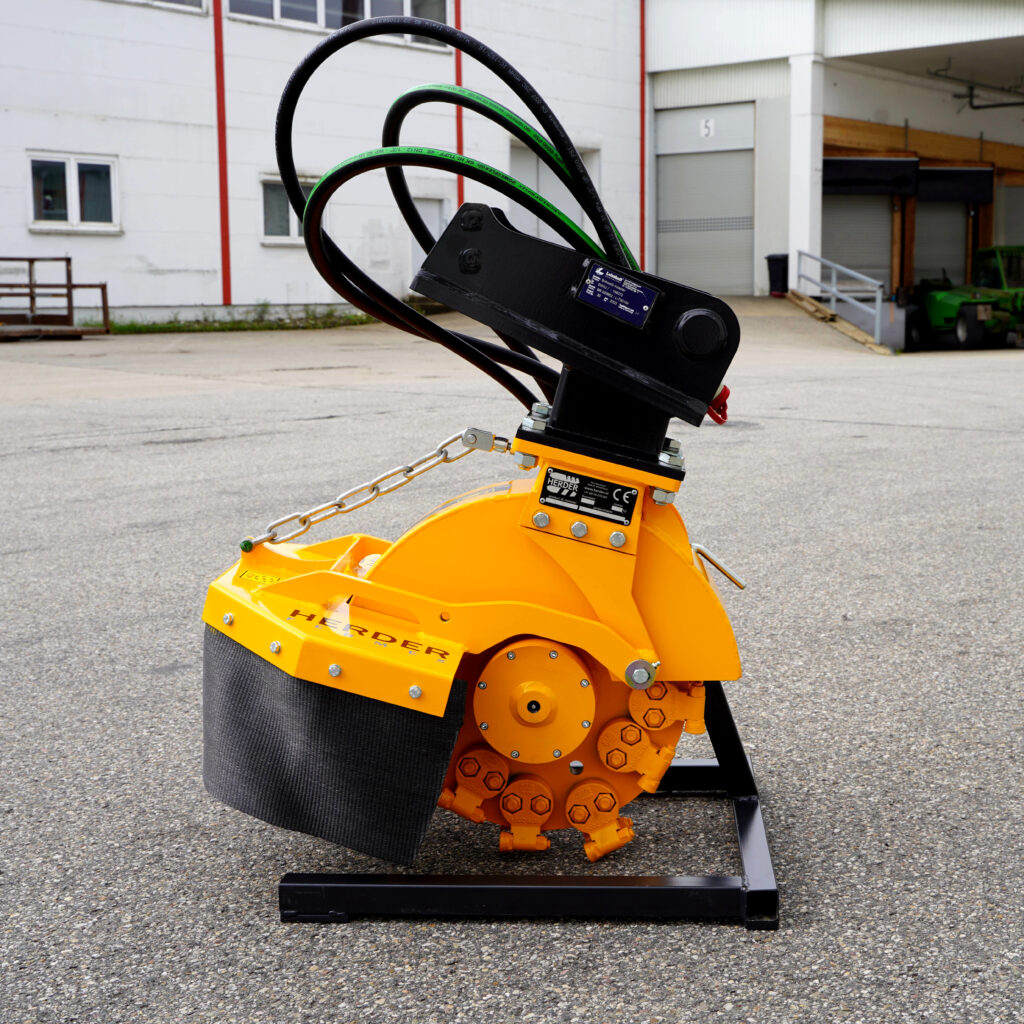 Ein gelb-schwarzer Herder-Stubbefräse SCE-410H-2 steht vor einer Industriehalle, mit massiven Fräszähnen, Hydraulikschläuchen und Ketten.