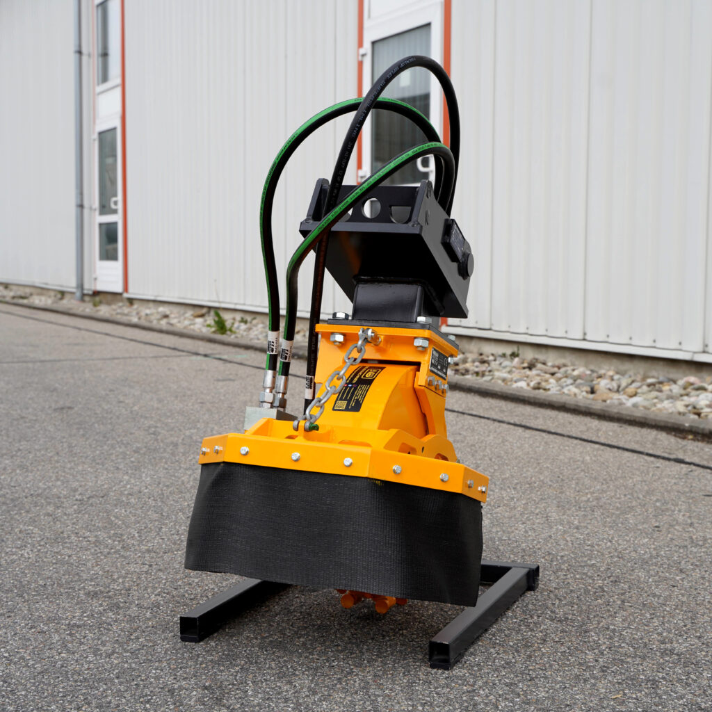 Ein gelb-schwarzer Stubbenfräsen-Aufsatz mit Hydraulikschläuchen steht vor einer weißen Industriehalle auf grauem Asphalt.