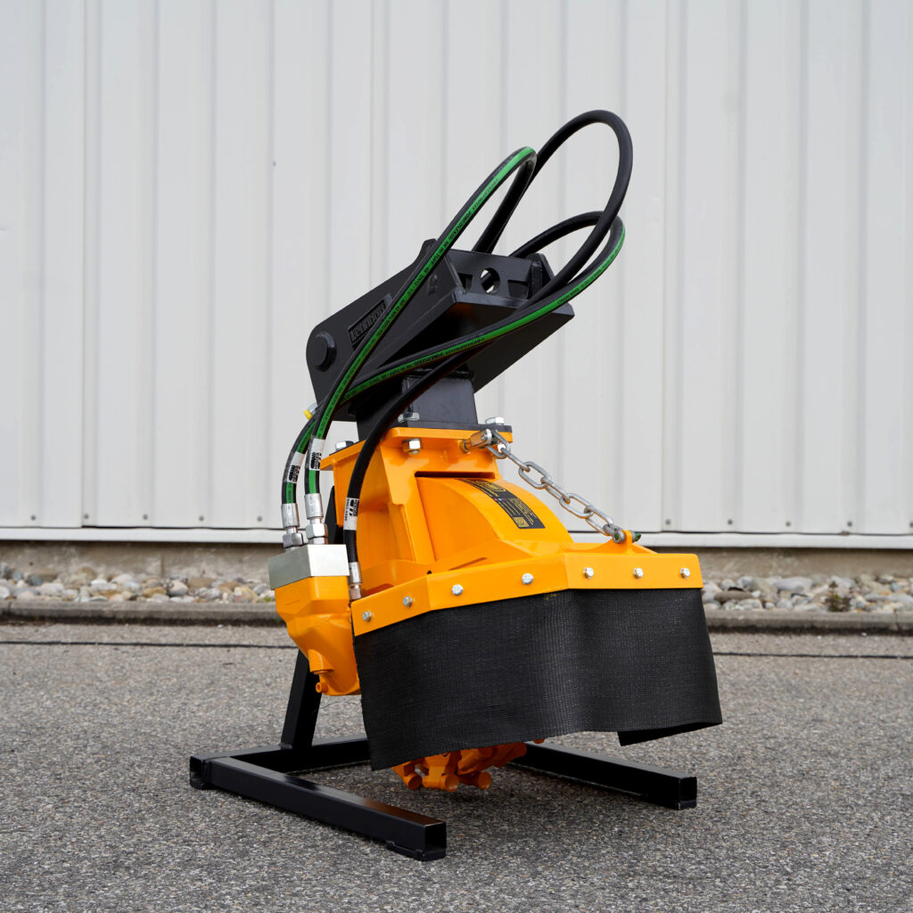 Ein gelb-schwarzer Stubbenfräse SCE-410H-4 mit Hydraulikschläuchen, Kettenzug und Stahlrahmen steht vor einer hellen Metallwand.