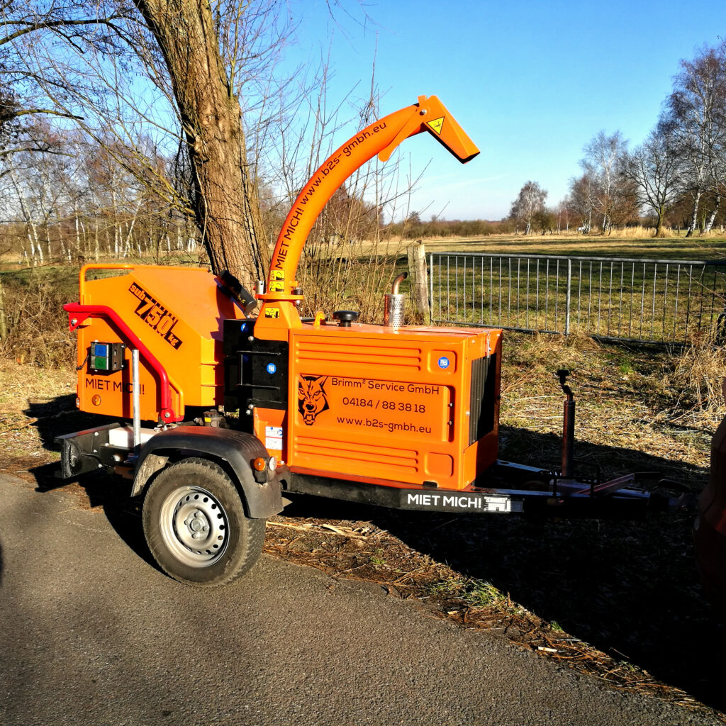 Ein orangefarbener Timberwolf-Holzhäcksler steht neben einem Baum auf einer Wiese, mit gebogenem Auswurftrichter, Anhängerfahrwerk und blauem Himmel.