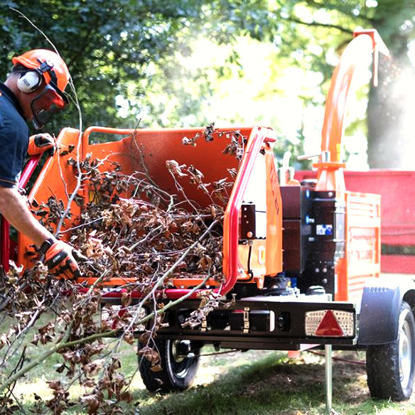 Ein orangefarbener Timberwolf TW230DHB verarbeitet dicke Äste, während ein Arbeiter mit Schutzhelm und Gehörschutz Zweige in den Einzug schiebt.