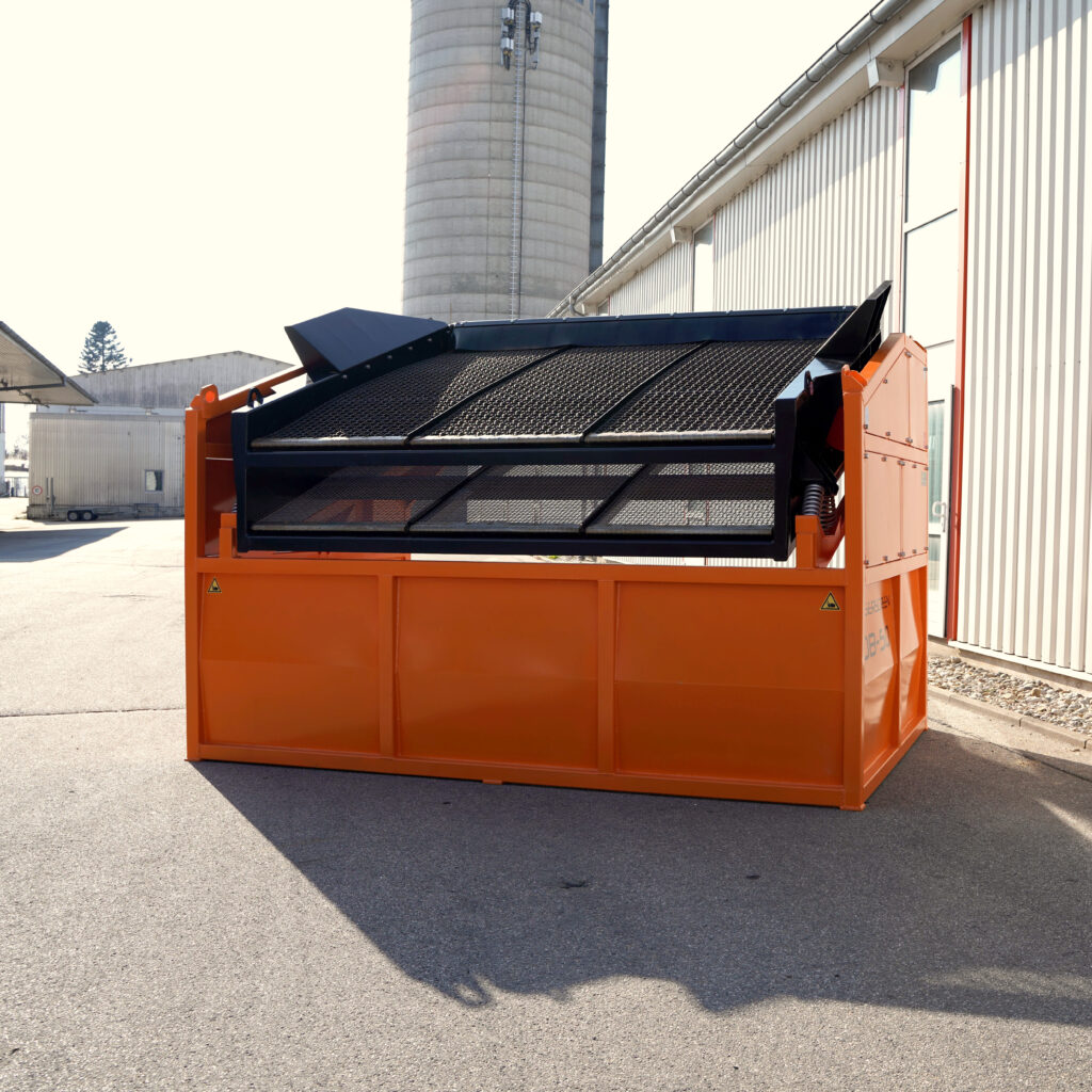 Orange-schwarze Traserscreen DB-50-1 Siebanlage mit Gitterdeck und großen Seitenwänden steht vor einer hellen Industriehalle neben einem Siloturm.