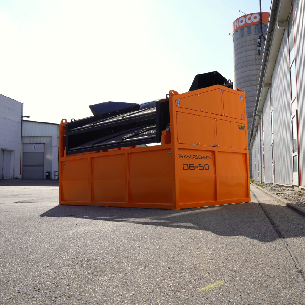 Großer orangefarbener Traserscreen DB-50 mit schwarzem Siebmodul steht zwischen Industriegebäuden unter hellem Himmel auf dem Betriebshof.