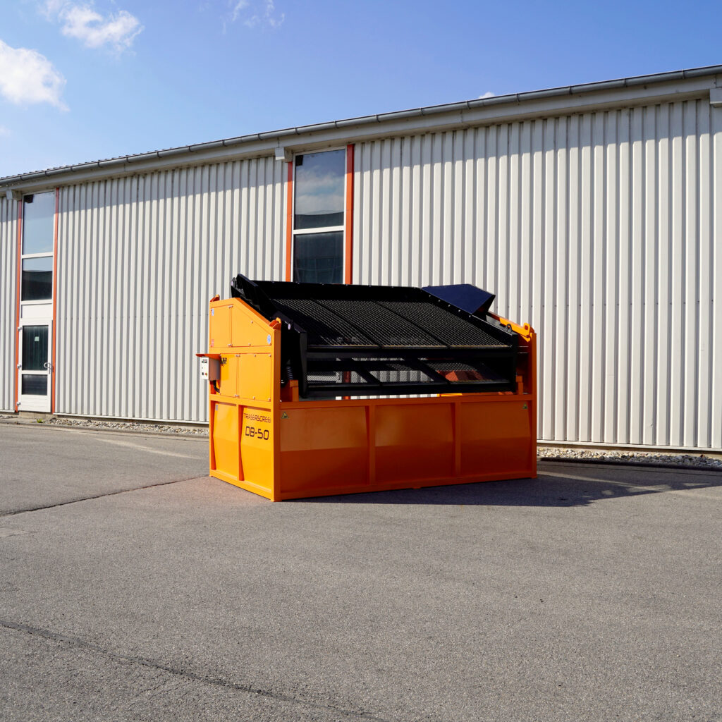 Der orange-schwarze Traserscreen DB-50 steht vor einer hellen Industriehalle unter blauem Himmel mit leichter Bewölkung.