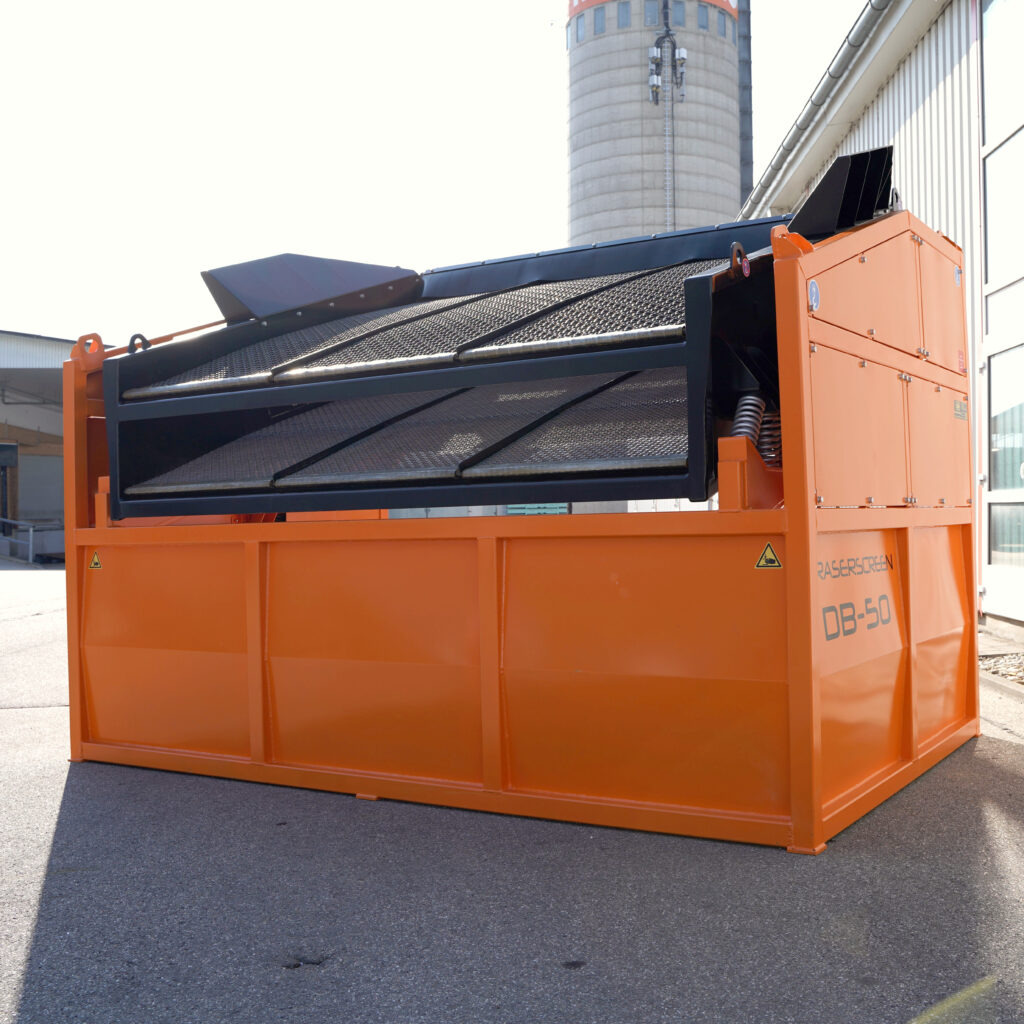 Vor einer Industriehalle steht ein orange-schwarzer Traserscreen DB-50-7 mit schräger Siebfläche und Metallgitter unter hellem Himmel im Freien.
