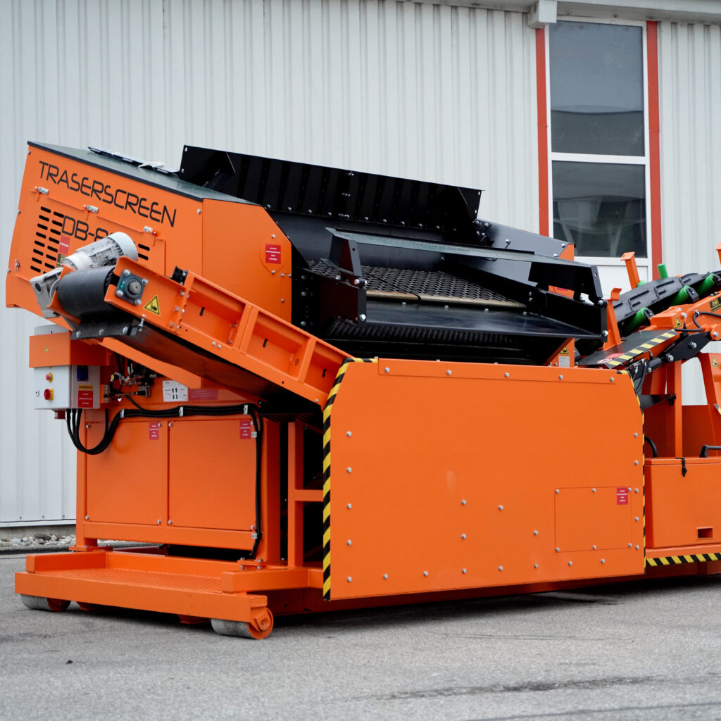 Ein orange-schwarzer Traserscreen DB-80-5 mit Förderbändern und Siebdecks steht vor einer hellen Hallenwand mit schmalem Fenster.