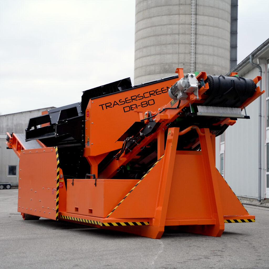 Ein orange-schwarzer Traserscreen DB-80 steht vor Industriehallen, mit großem Trommelmodul und sichtbaren gelb-schwarzen Warnstreifen.