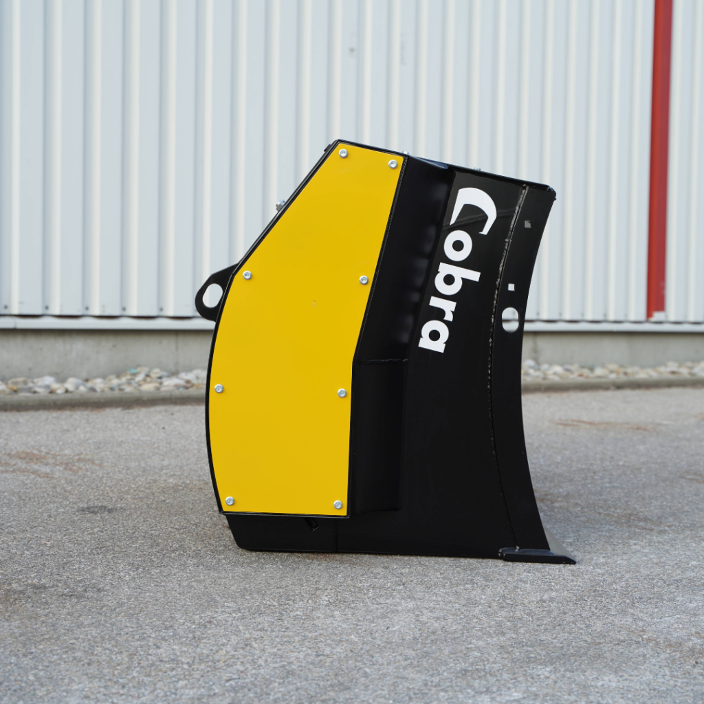 Ein schwarz-gelbes Cobra-Anbaugerät steht auf Asphalt vor einer hellen Metallwand und zeigt seitlich das markante, gebogene Gehäuse mit Logo.