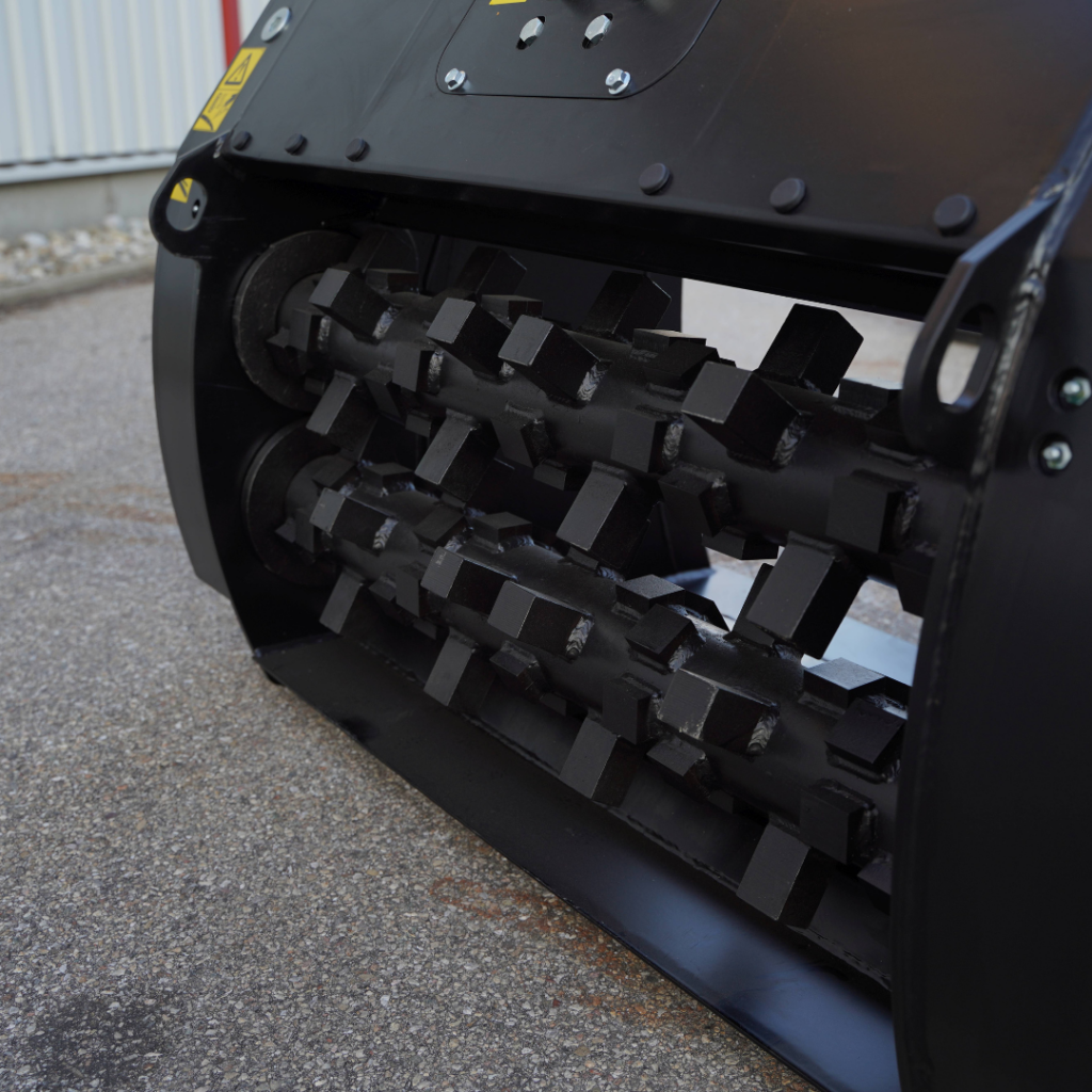 Ein schwarzer Brecherkopf mit zwei massiven Walzen und aufgeschweißten Zähnen liegt im Vordergrund auf dem Asphalt vor einer Industriehalle.