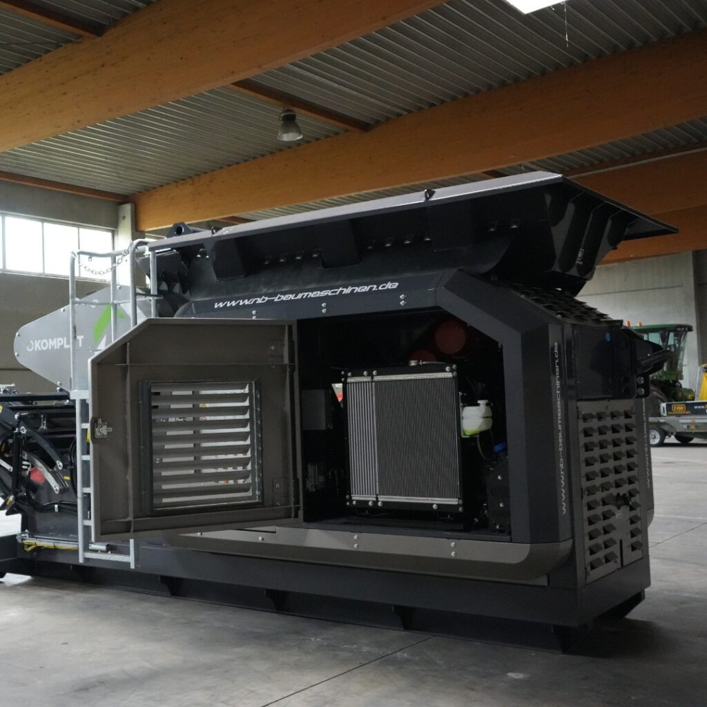 Große schwarze Recyclingmaschine in einer Halle mit geöffneten Wartungsklappen, sichtbarem Kühlmodul und massiver Stahlkonstruktion unter Holzbalken.