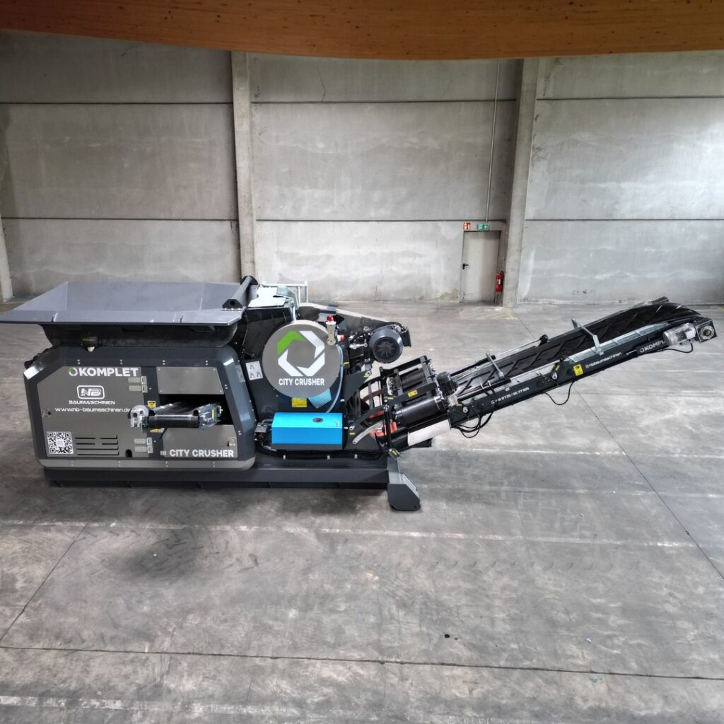 In einer großen Halle steht der graue Komplet City Crusher mit schwarzem Förderband und blauem Aggregat auf glattem Betonboden vor einer Betonwand.