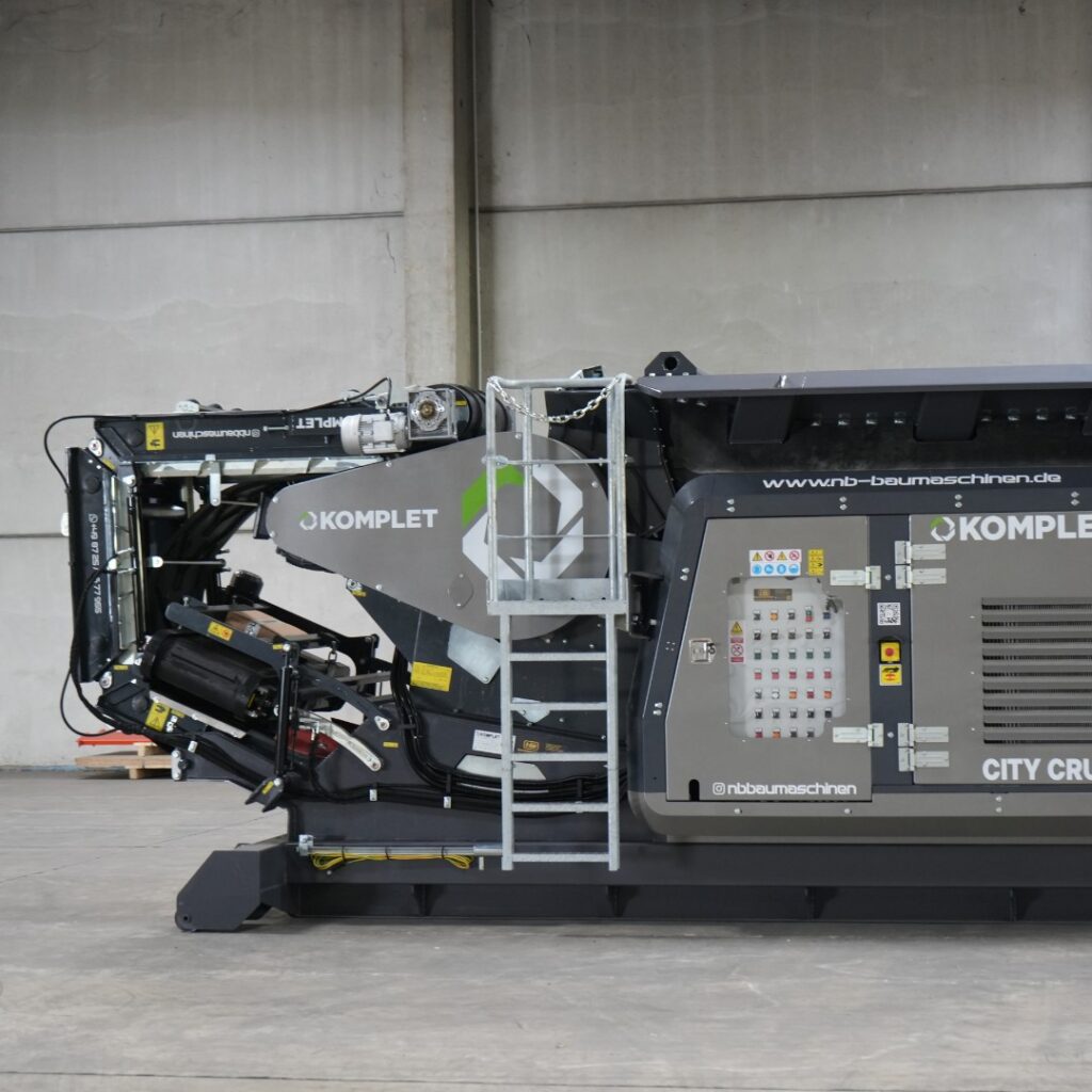Die mobile Komplet City Crusher steht in einer Halle, mit grauer Verkleidung, schwarzem Förderband, Leiter, Bedienfeld und gelbem Not-Aus.