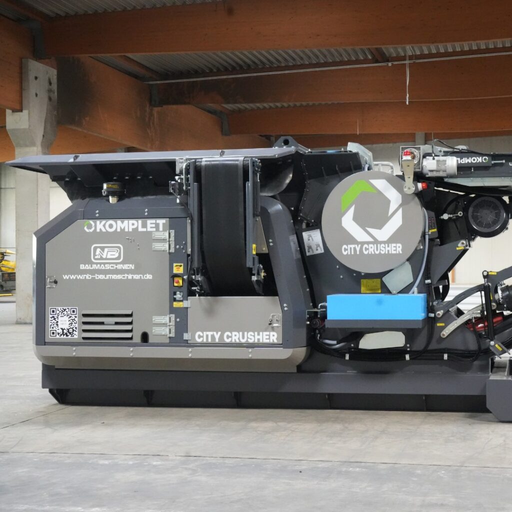 Der graue Komplet City Crusher steht in einer Halle, mit sichtbarer Technik, Förderband und großem Rundgehäuse in klarer Seitenansicht.