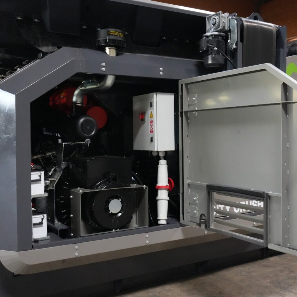 Eine große Recyclingmaschine mit offenem Seitentor zeigt Motoren, Rohre, Steuerkasten und schwarze sowie graue Gehäuseteile in einer Werkhalle.